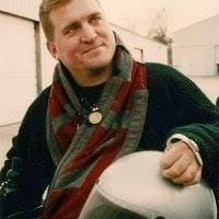 Steve Foelsch's head is tilted to the side with a smirk on his face. Steve has fair skin, blonde hair and is wearing a red and green scarf with a black sweater.