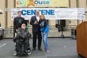 4 people smile on an outdoor stage. One man uses a wheelchair, another man stand behind him, a man receiving a glass award statue stands arm-in-arm with a woman. Signs behind them say Centene Charitable Foundation. Tshirts in the photo read ADA 25