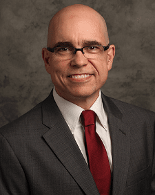 Michael Reese poses for a headshot. He is wearing black glasses, a grey suit and a red satin tie.