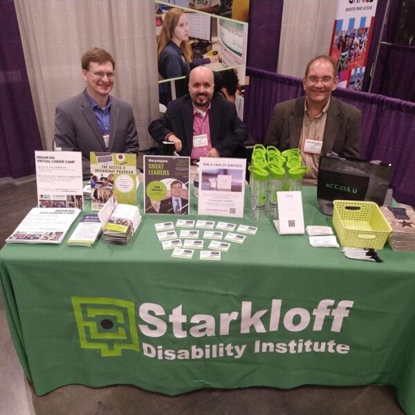 Jason, Brian and Scott sit behind the Starkloff event table. The table is covered in a green tablecloth and Starkloff swag is placed on top for attendees.