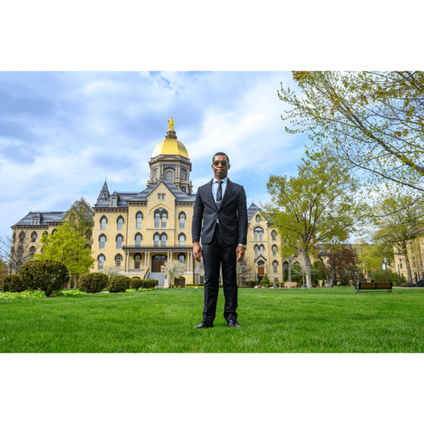 Justice is standing on grass in front of a domed cathedral. He is wearing a navy suit and has on grey sunglasses.