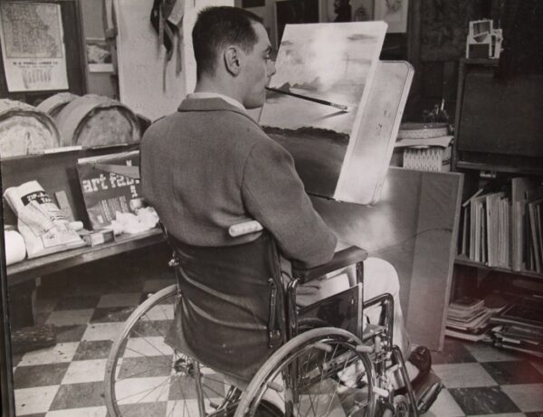 Black and white photo of Max Starkloff as a young man painting on a canvas. He is in a sports jacket and seated in his manual wheelchair, gripping the long paintbrush with his teeth, painting what looks to be a beautiful landscape.