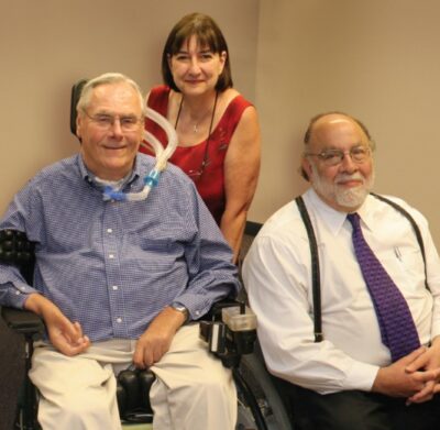 Photo of Max and Colleen Starkloff and David Newburger. Max and David are both seated in their wheelchairs and Max’s vent tube is visible. Colleen stands between them as all three smile at the camera.