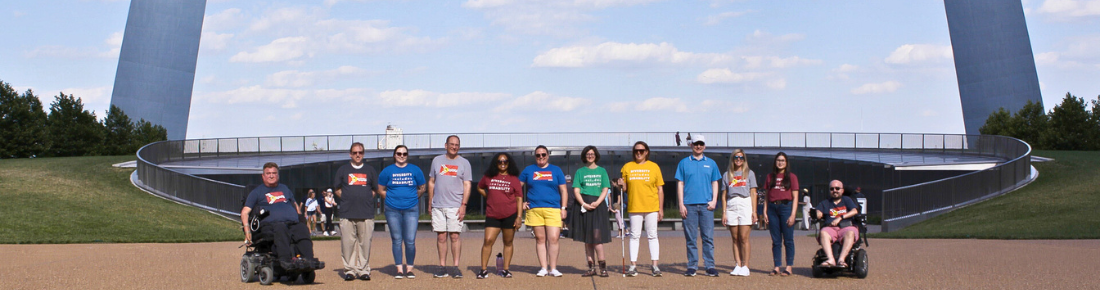 Starkloff staff and community members in colorful disability pride tshirts smiling in a line under the Gateway Arch