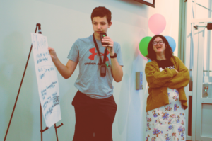 Student gives presentation while talking into an FM transmitter. A staff person stands in the background listening, with a big smile, standing near balloons.