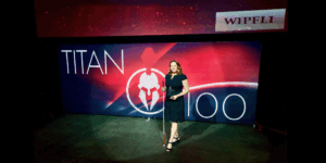 Lori Becker on a dramatically lit stage, holding her white cane and a glass award. The backdrop is red and black with a large Titan 100 logo, and Wipfli logo.