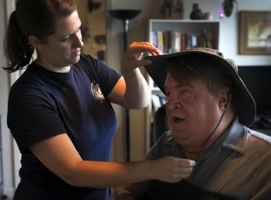 Steve seated in his power wheelchair, talking with his PCA who is assisting him in getting ready to head to work. She is adjusting his hat for sun protection.