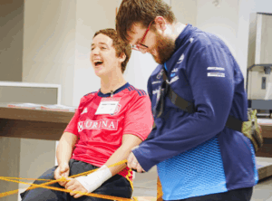 Two students laugh while holding parts of a large yarn web.