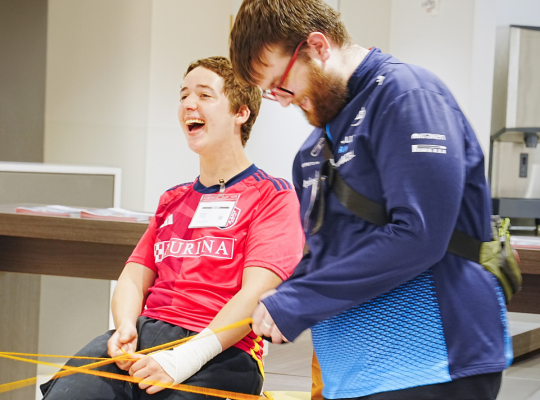 Two students laugh while holding parts of a large yarn web.