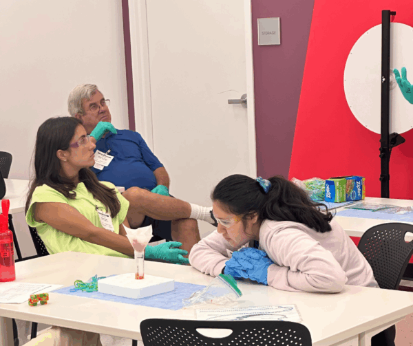 In a science learning lab, a student observes an experiment as Camp counselor and driver listen to an instructor.