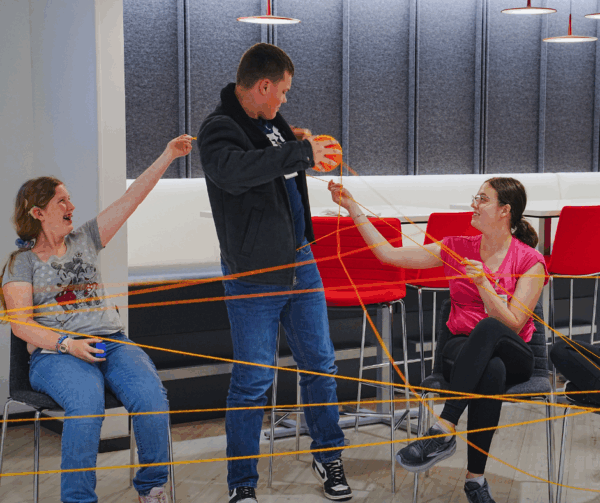 Three students laugh while holding parts of a large yarn web.
