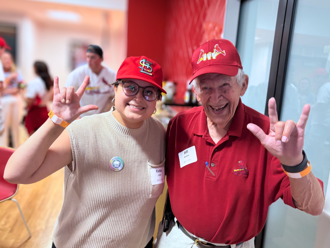 Two people in Cardinals Disability Pride Night hats smile with ASL signs signifying Deaf pride.