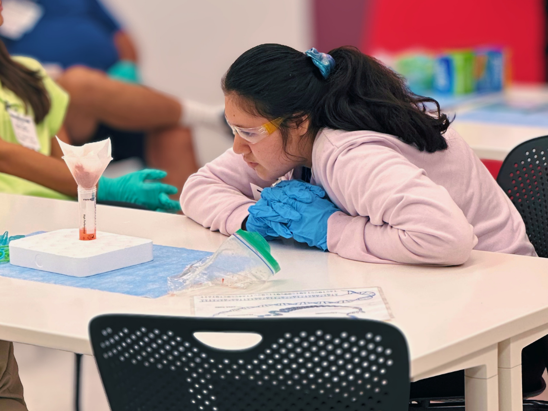 Student wearing protective glasses and gloves examines graduated cylinder.