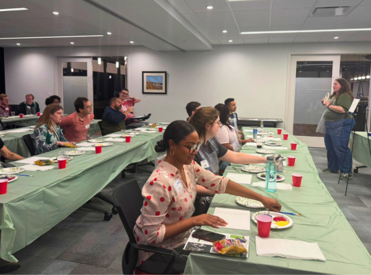 Paint and Sip – Mack teaching group Young adults at rows of tables in a conference room work with paints on their individual canvases, guided by an instructor.