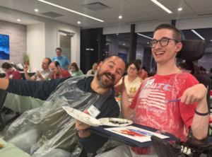 Two people smiling during the painting process. One is a power wheelchair user.
