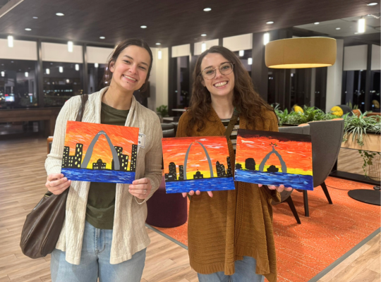 Paint and Sip – finished canvases Smiling people show three similar small paintings of the St. Louis skyline, all featuring the Arch and a fiery orange sunset.