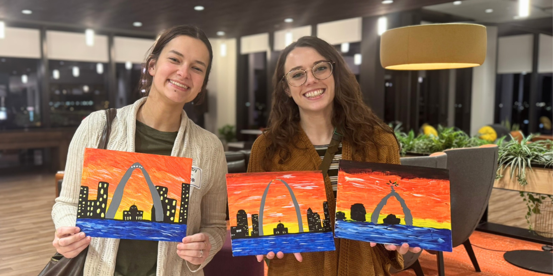 Smiling people show three similar small paintings of the St. Louis skyline, all featuring the Arch and a fiery orange sunset.