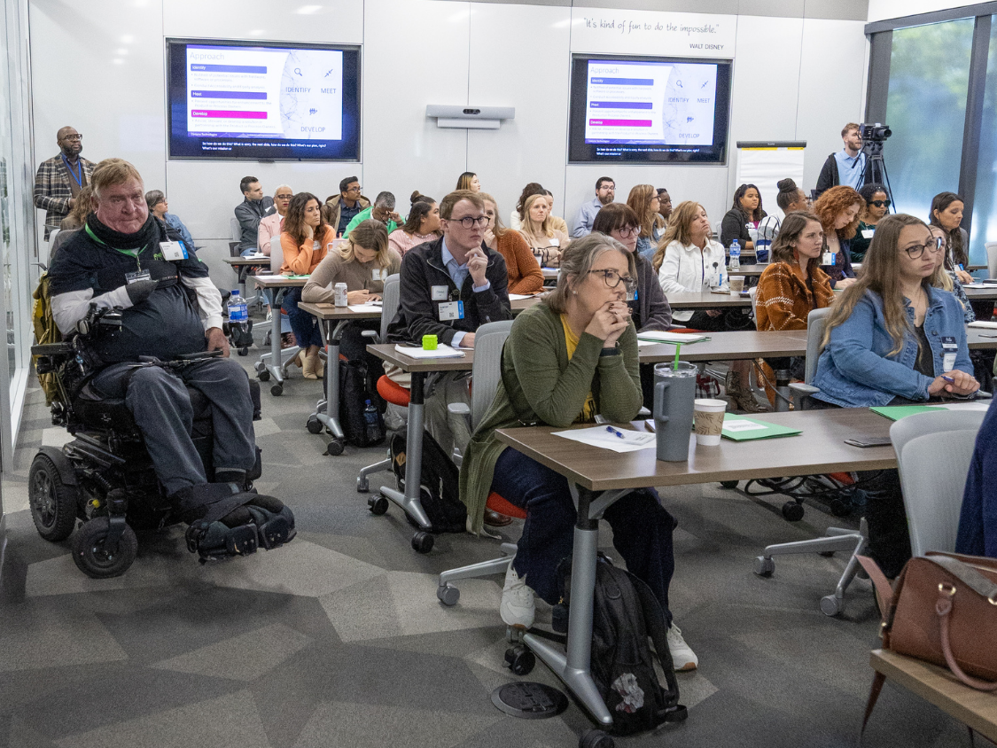 A conference room full of professionals with and without disabilities taking notes during a presentation.