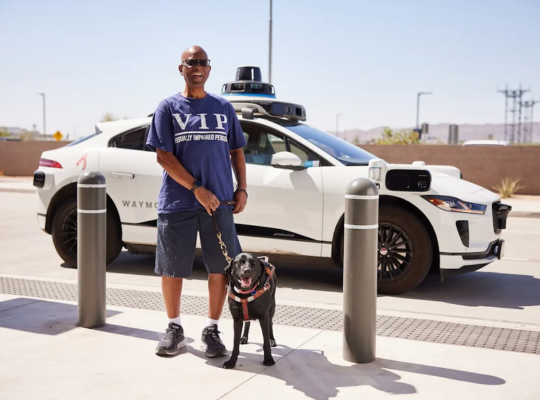 Sheldon and his guide dog, Nora, both stand smiling toward the camera in front of a Waymo autonomous vehicle.