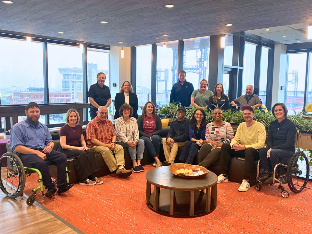 16 people smile as a group. Some have obvious disabilities, including three wheelchair users.