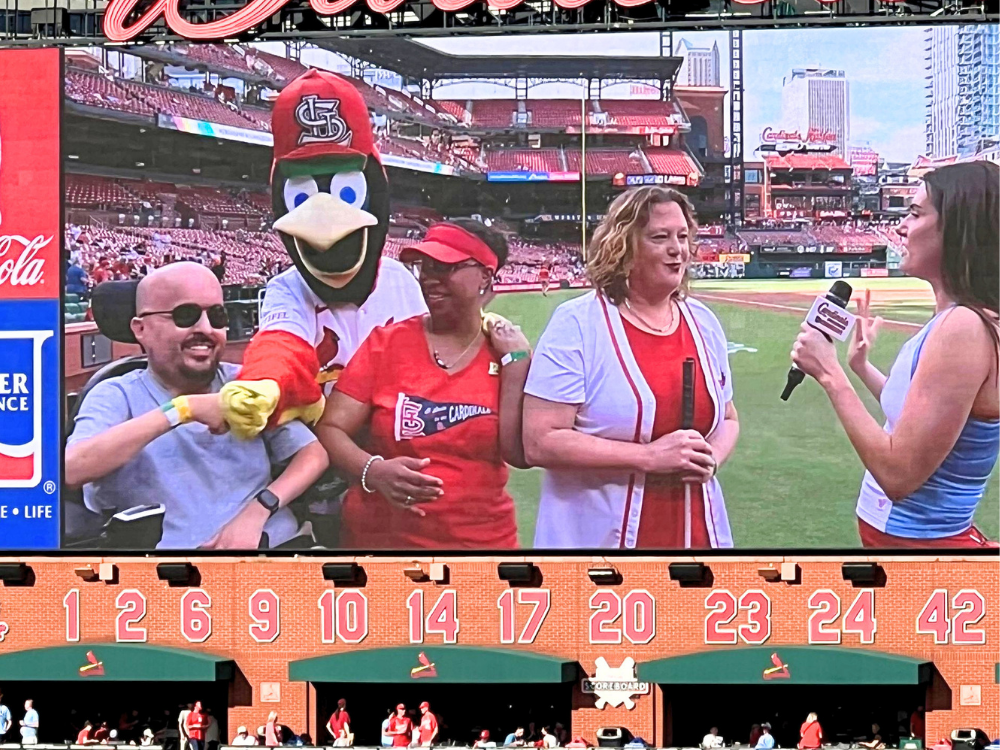 Scene of the big screen at Busch Stadium showing 3 guests for an interview and fist bumping Fredbird. One person is a power wheelchair user and another holds a white cane.