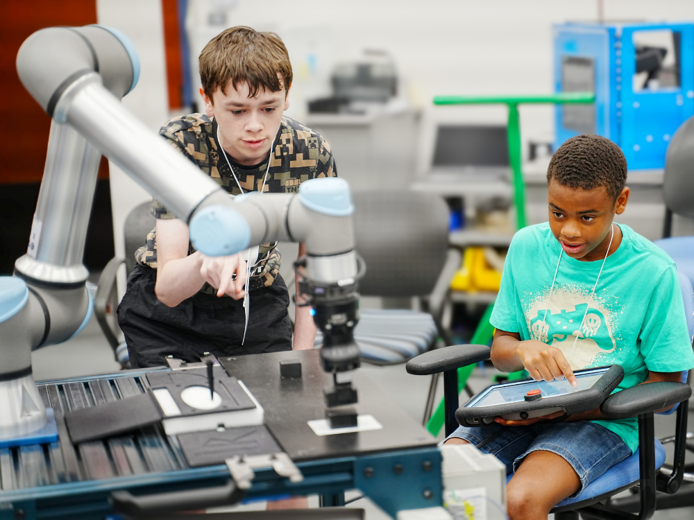 Two tween boys operate a robotic arm in a classroom lab.