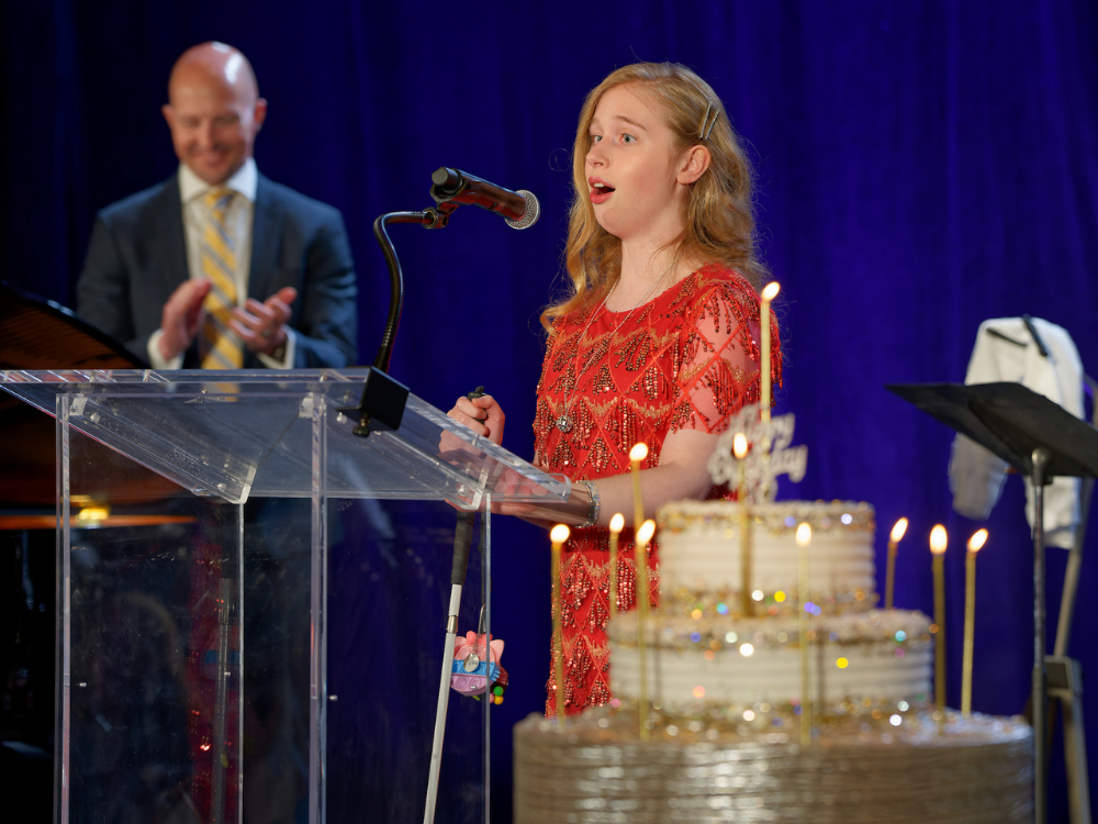 Birthday song led by Holly. Holly Conor, a teenager holding a white cane, sings onstage in a beautiful red beaded dress near a large birthday cake.