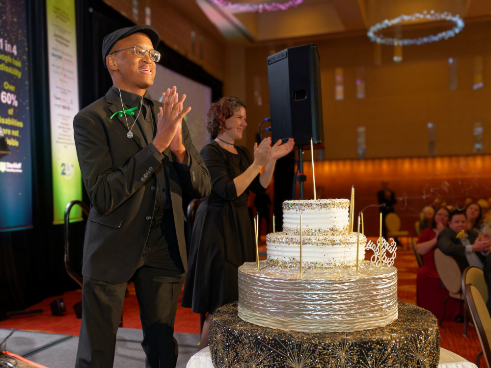 Christopher-birthday-candles A young professional in a black suit with a mini disco ball necklace smiles and claps with ballroom audience while standing on stage with a 5 tier birthday cake, candles extinguished.