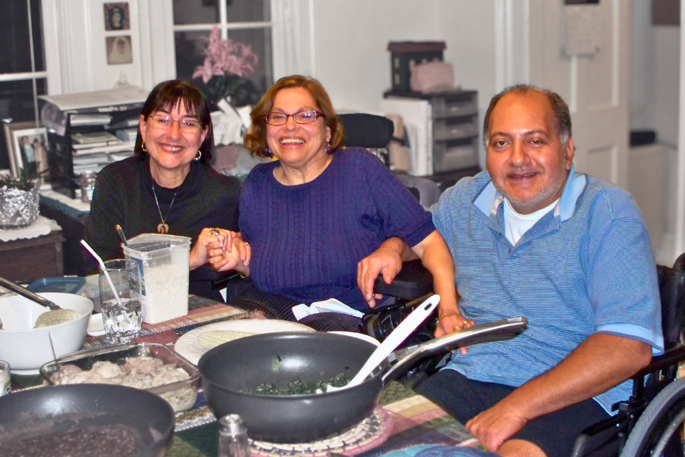 Colleen-Starkloff_Judy-Heumann_Jorge-Pineda JColleen Starkloff, Judy Heumann, and Jorge Pineda smile brightly, shoulder to shoulder with hands and arms woven together, at Judy and Jorge’s kitchen table with a delicious home-cooked meal on the table in front of them.
