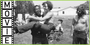 Movie: Two men laugh together as one carries the other in a very 1970s scene with several young people hanging out in a large grassy area, many of them seated in manual wheelchairs.