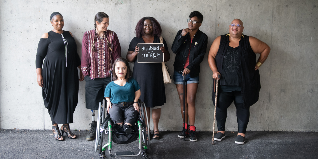 Six disabled adults of diverse ages, skin tones, hair styles, physical size, gender identities, and support needs smile together as friendly group. One person holds a sign that reads Disabled and Here in joyful lettering.