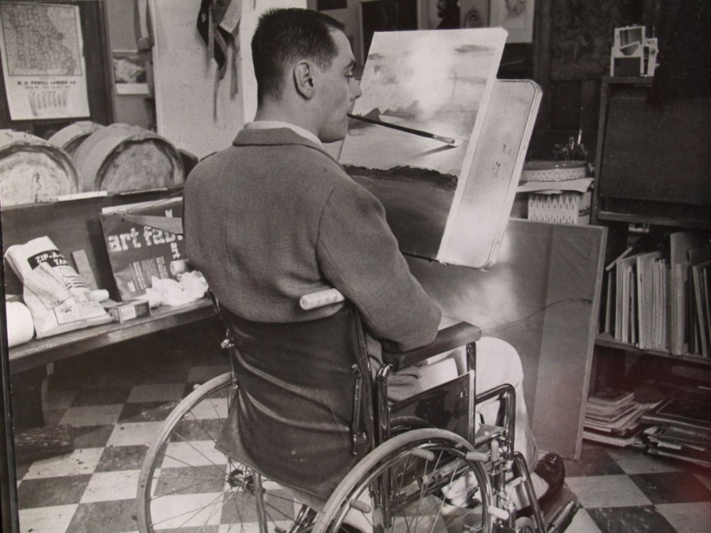 Max Starkloff painting Max Starkloff as a young man seated in his manual wheelchair and painting a landscape, holding the brush with his mouth.