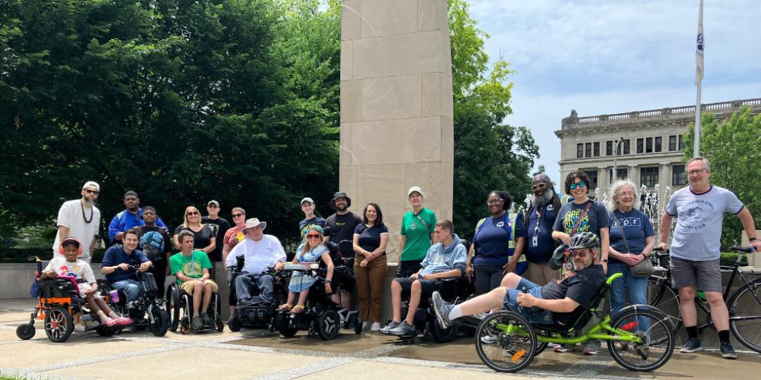 A large group pedestrians smile together on a sunny day at Soldiers Memorial. Some people are using wheelchairs or scooters and a couple have bikes.