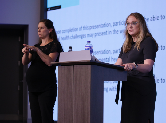 Person giving presentation with a large screen behind them, a podium, and an ASL interpreter.
