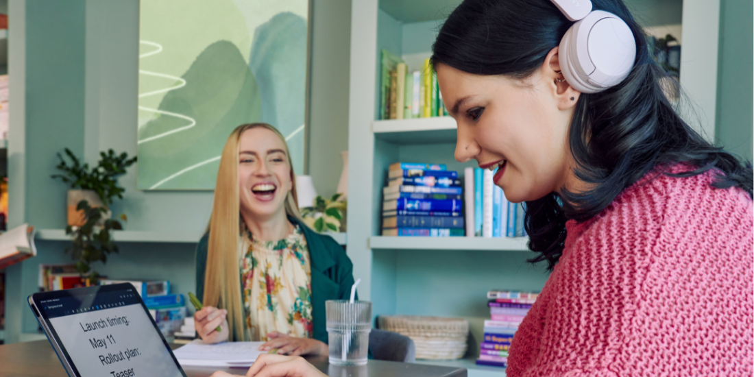 A young pale white blind woman is sitting at a table, typing on a keyboard connected to a tablet computer in a modern coworking office space. Visible on her tablet is a document in large print that reads "Launch timing: May 11. Rollout plan: Teaser. Drumbeat.\" She is smiling as she types. Across from her at the table, a young white woman with partially paralyzed arms and hands laughs with the blind woman as she holds a pen to a notebook in front of her. A glass of water with a straw in it is nearby. The woman with partially paralyzed arms and hands has long straight blonde hair and is wearing a floral dress and green blazer. The blind woman has shiny black hair styled in waves and is wearing over the ear headphones that partially cover one ear and a chunky knitted pink sweater. The coworking office space is painted a warm mint green and features a large bookcase with several books, plants, and a large abstract art piece.