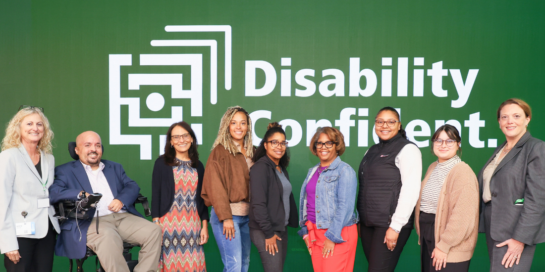 A group of professionals, some with obvious disabilities, smile together in front of a large screen featuring Stakrloff's Disability Confident tagline.