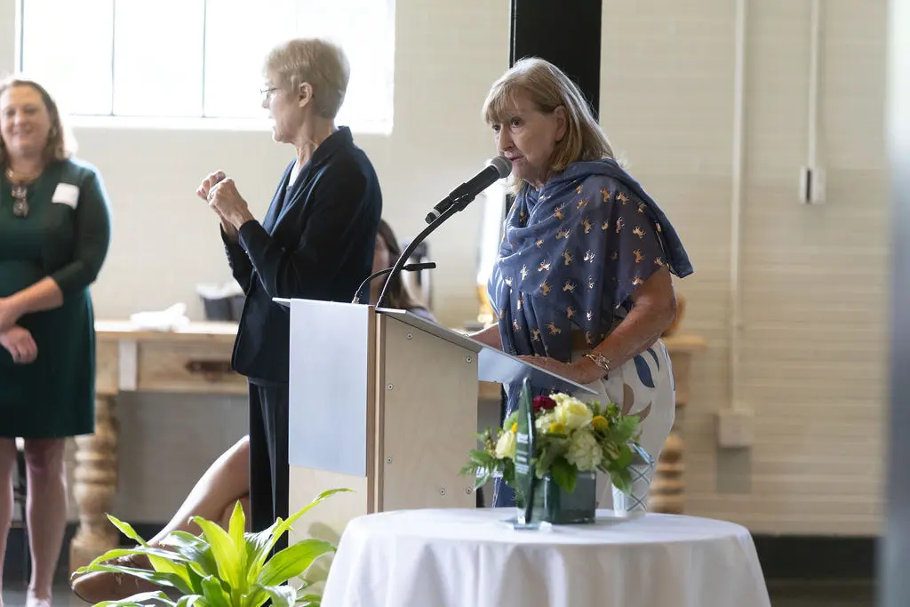 Colleen Starkloff makes remarks at a podium with and ASL interpreter nearby. The award recognizing Ray Hartmann is on a table with flowers on her other side.